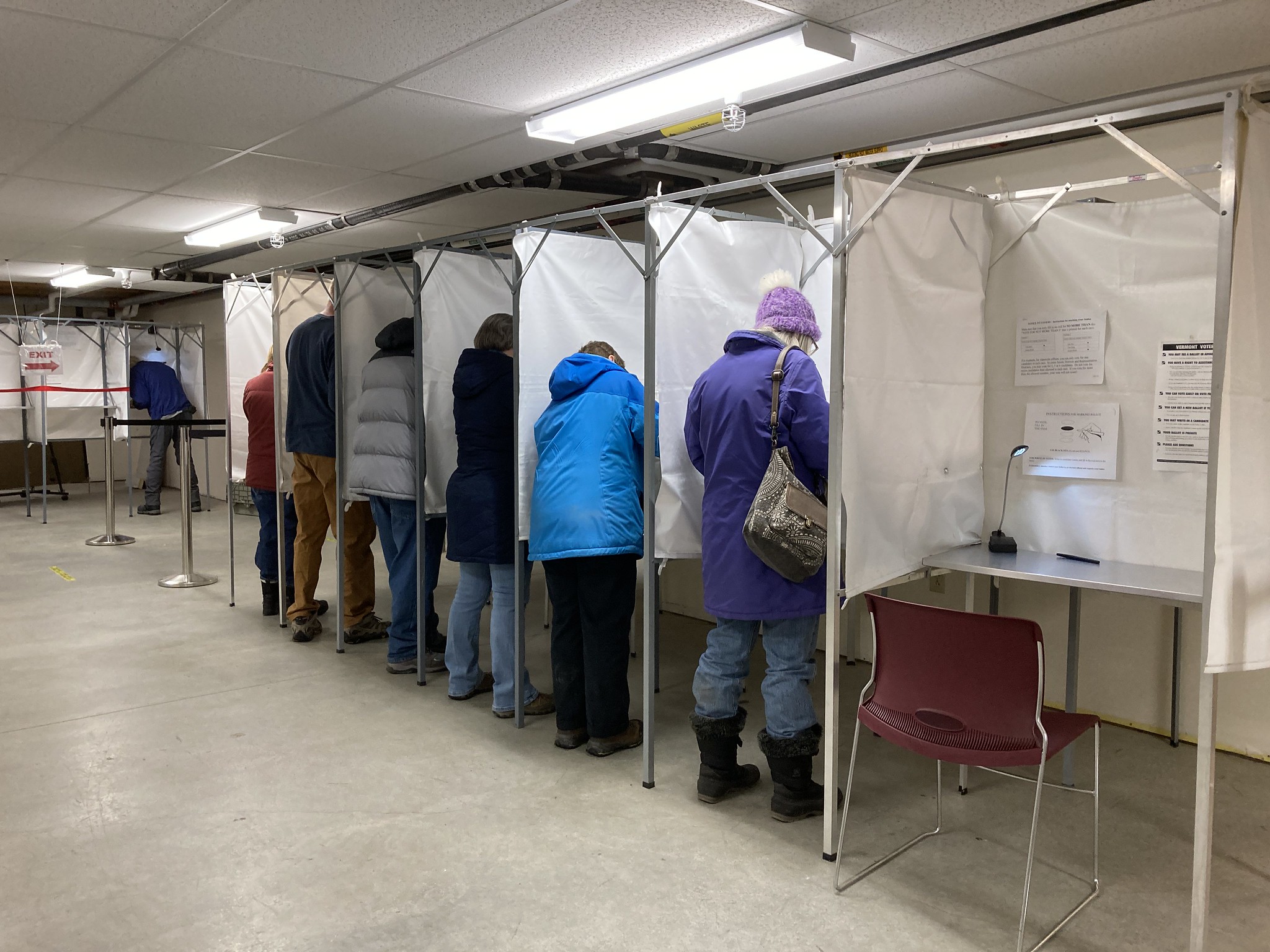 A row of people wearing winter gear voting in booths in the basement of the town hall