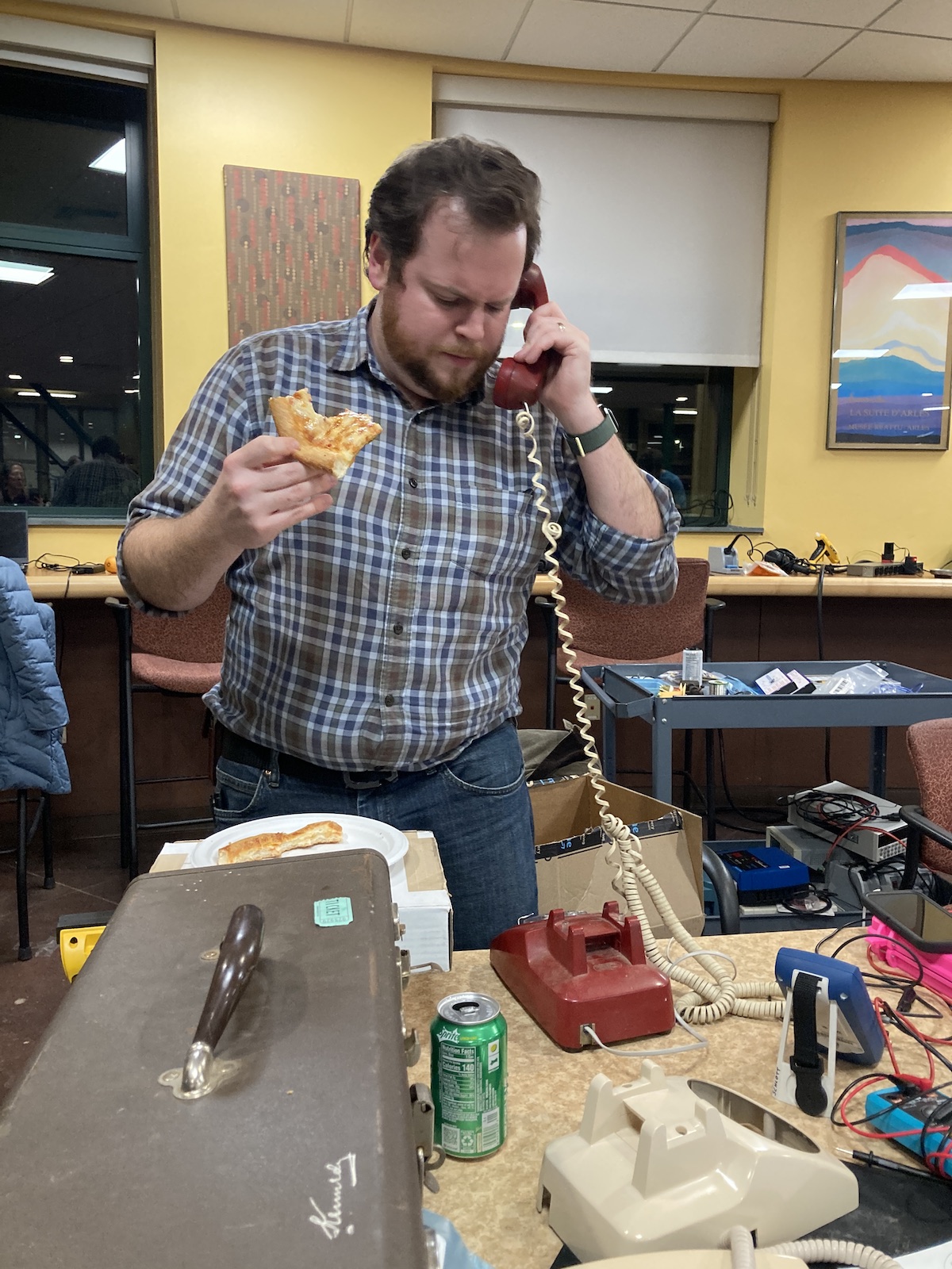 a man holds a slice of pizza and talks into an old school telelphone
