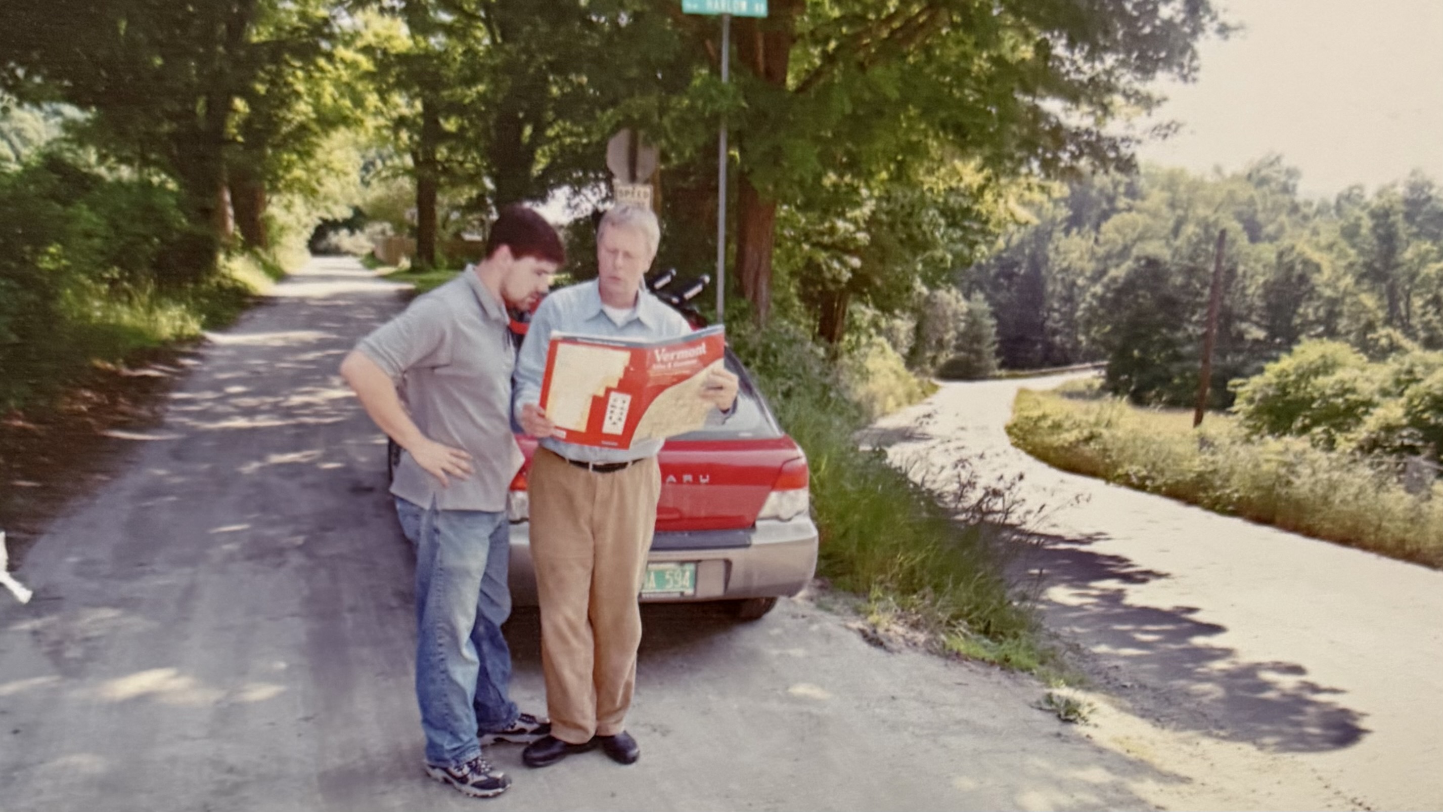 two men standing on a dirt road beside a car 